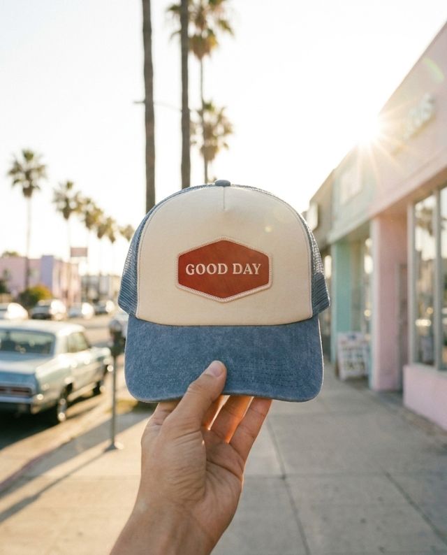 AI variant: point-of-view hand holding the GOOD DAY cap on a palm-lined street